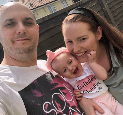 A photo of a couple beside a body of water. The woman is looking directly to camera.